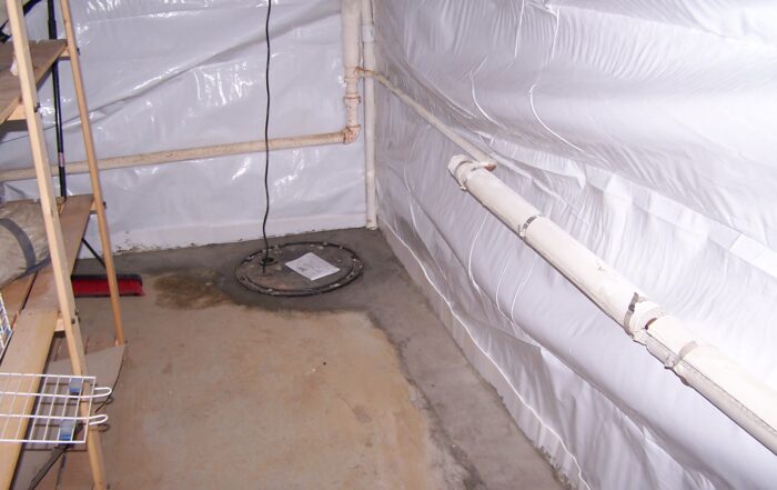 Basement corner with exposed white pipes and concrete floor, showing a sump pump cover. Shelves are on the left, broom nearby. It feels utilitarian.