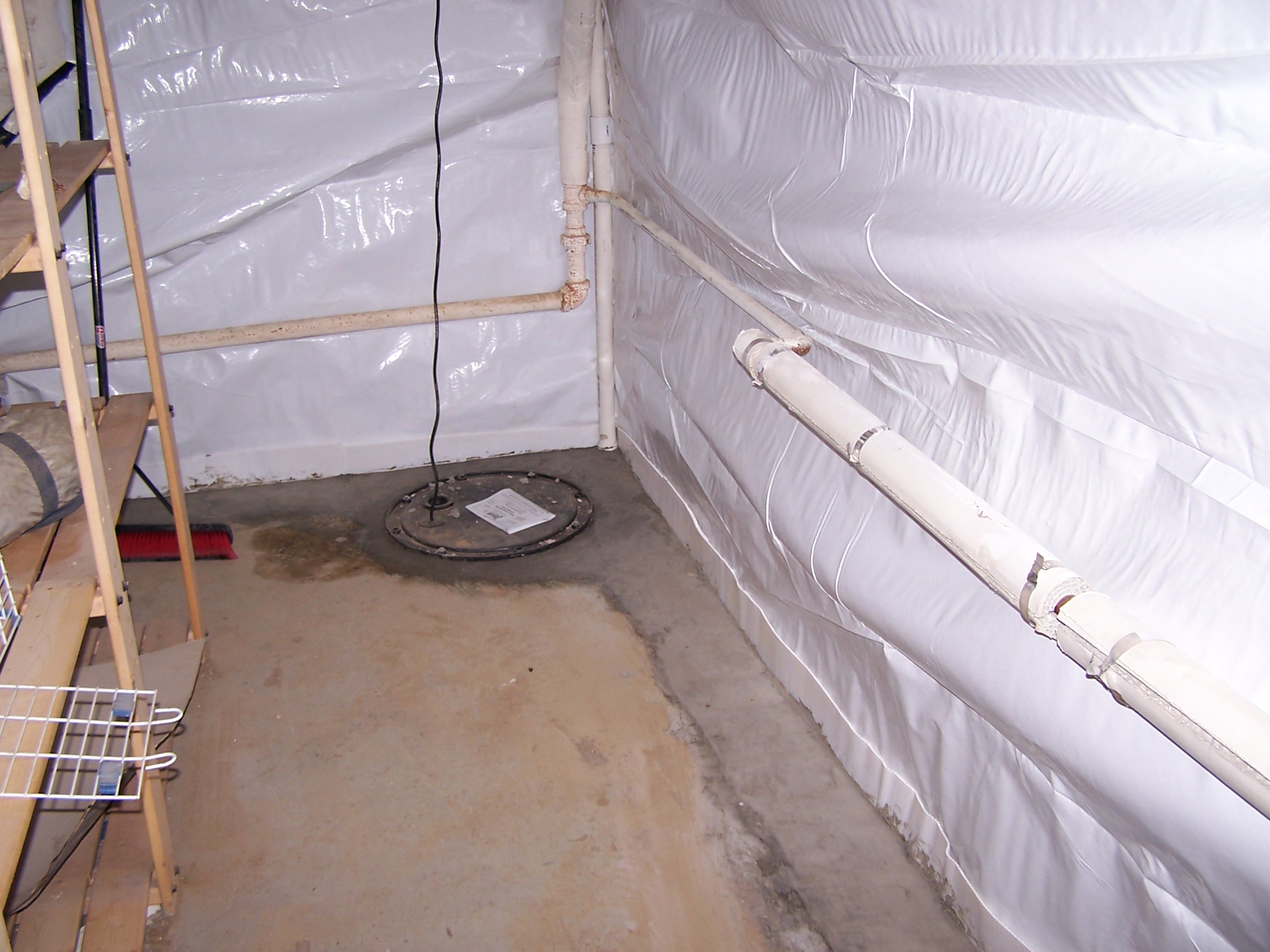 Basement corner with exposed white pipes and concrete floor, showing a sump pump cover. Shelves are on the left, broom nearby. It feels utilitarian.